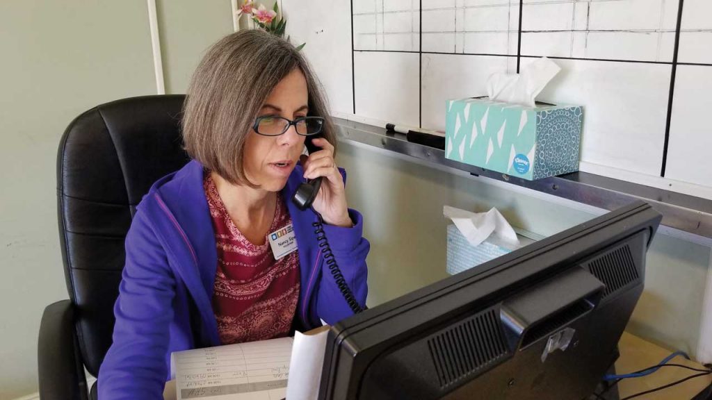 Nancy Sipe sits at desk with phone to ear looking at a computer monitor wearing a purple jacket and dark rimmed glasses.