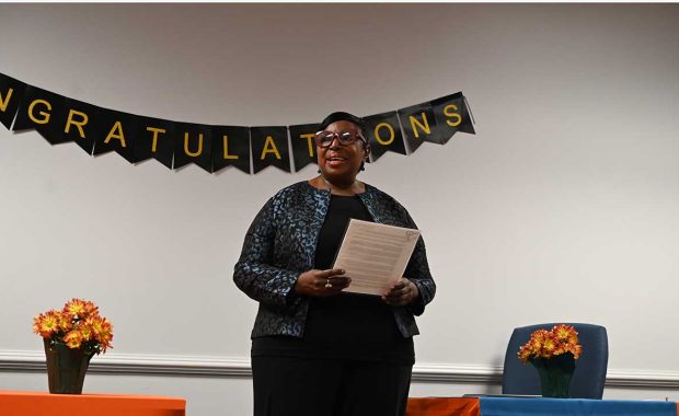 Deborah stands to speak holding her notes in a decorated conference room.